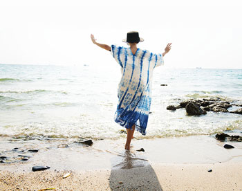 woman doing yoga tree pose in blue and white tie dye dress and sunhat on a beach